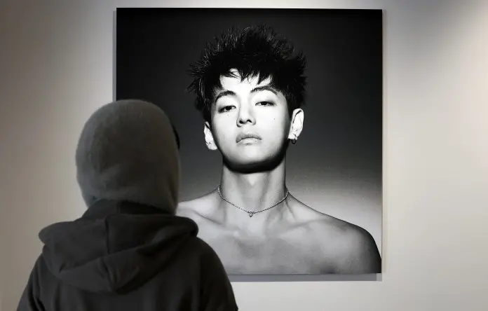 A person wearing a hooded coat stands in front of portrait of a V with a serious expression, short spiky hair, and earrings.