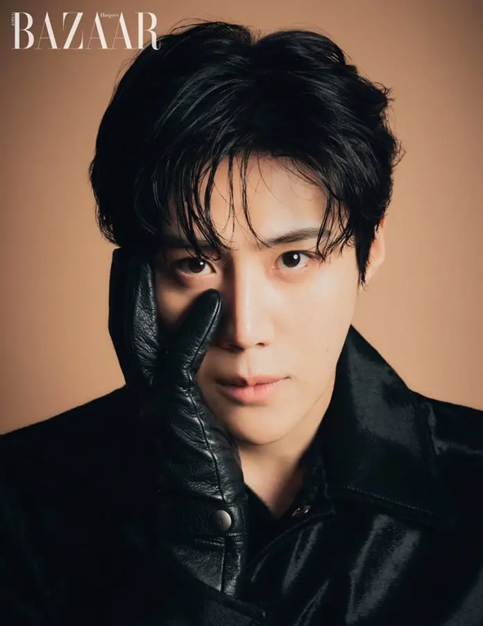 A close-up portrait of Kim Seon-ho with tousled black hair, wearing a black jacket, partially obscured by a black leather glove over one eye. The background is a soft, neutral color.