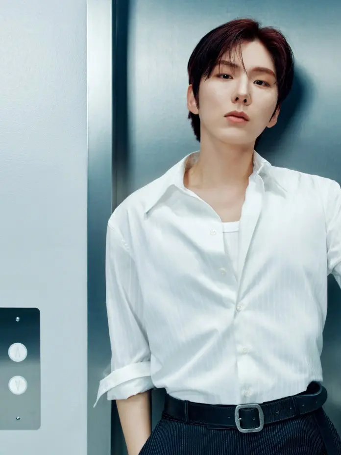 Kihyun standing in an elevator, wearing a white striped shirt with rolled-up sleeves and dark pants, looking directly at the camera.