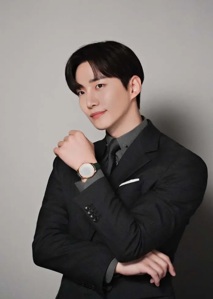 Lee Junho in a black suit posing confidently with a watch on his wrist, smiling slightly at the camera.