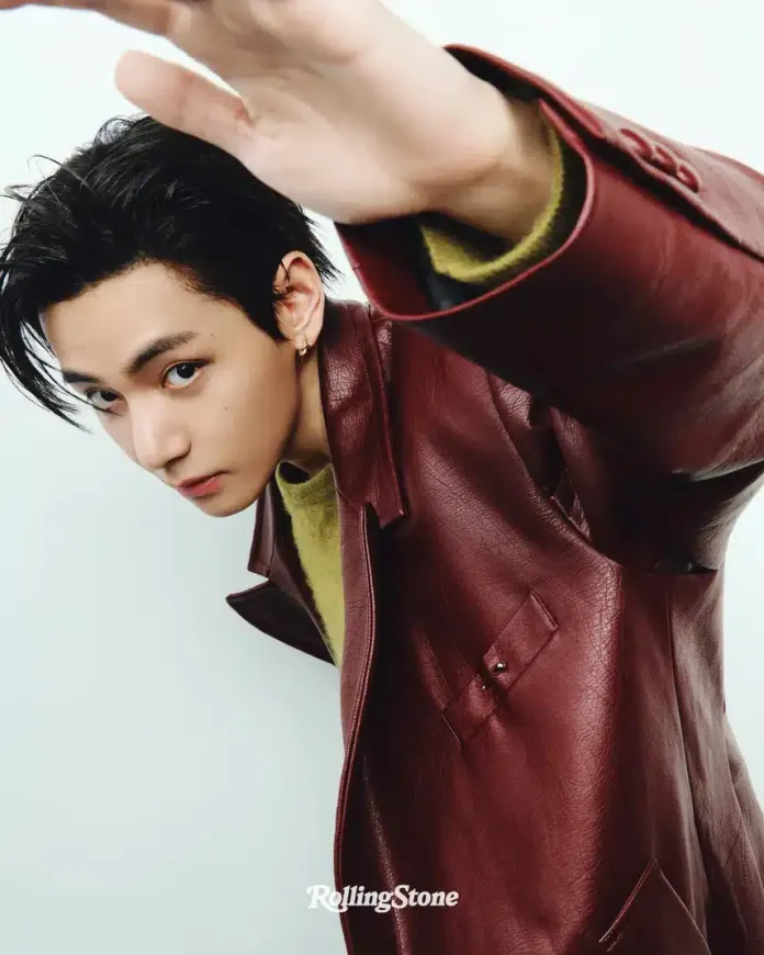 A close-up photo of BTS V with dark hair and piercing eyes, wearing a burgundy leather jacket over a green sweater, reaching out with his hand against a light background.