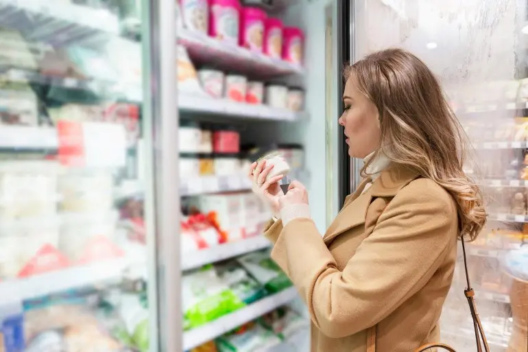 Woman buys ice cream in the store. Young beautiful blonde in a beige coat opened a refrigerator door in the supermarket. ; Shutterstock ID 2073038219; purchase_order: aj; job: ; client: ; other:
