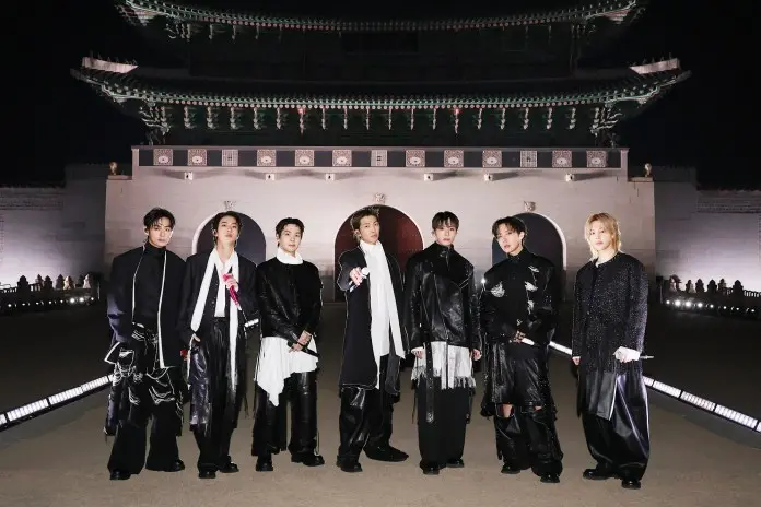 BTS dressed in stylish, dark clothing stand in front of a traditional Korean palace, illuminated by night.