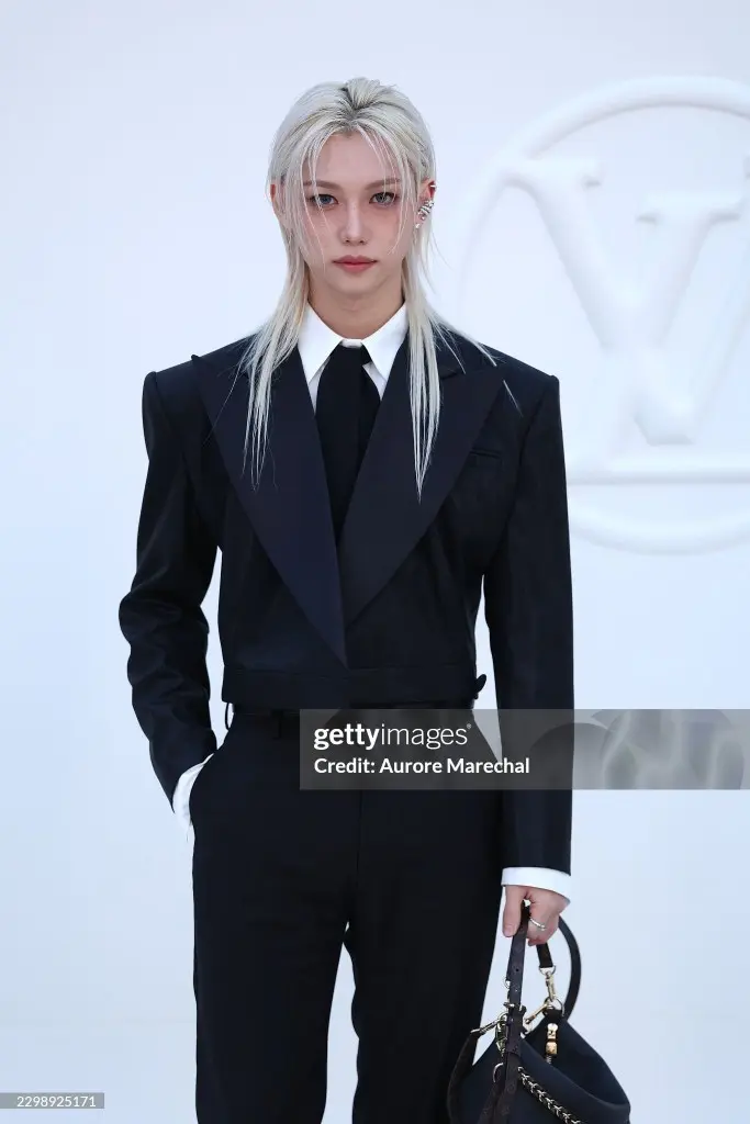 Stray Kids’ Felix wearing a stylish black suit with a white shirt and tie, standing confidently while holding a designer handbag.