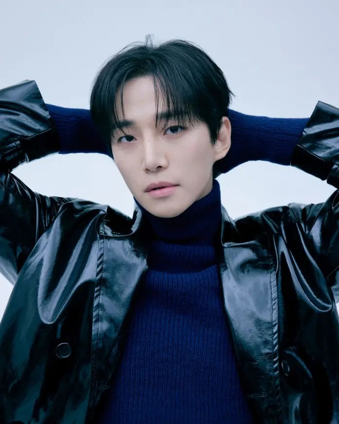 A close-up portrait of Lee Junho with dark hair wearing a glossy black jacket and a dark blue turtleneck, posing with hands behind their head against a light background.