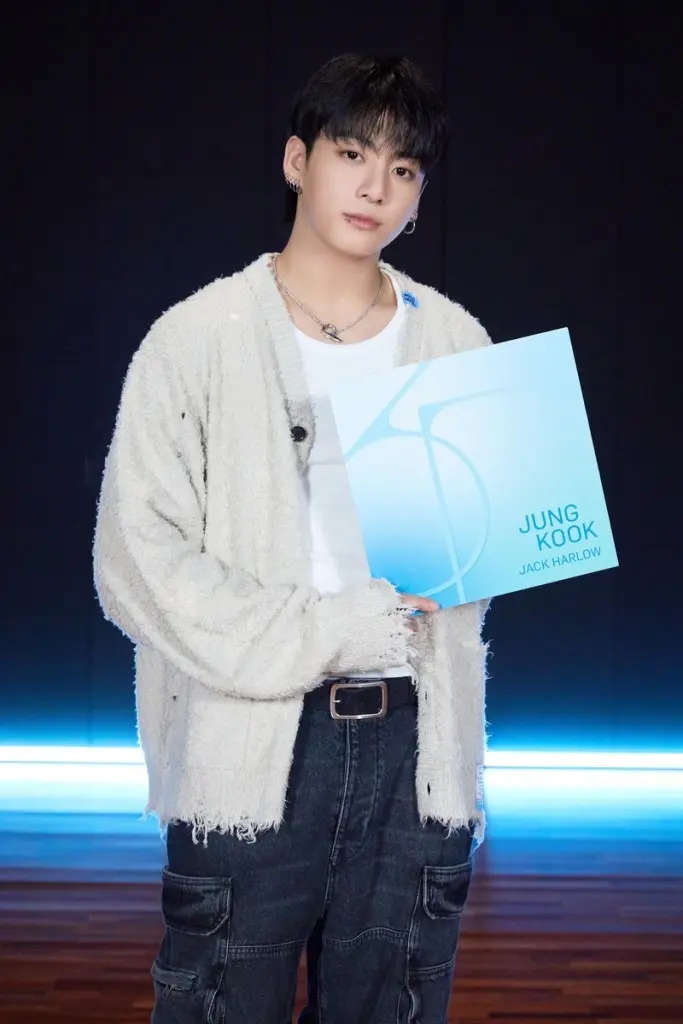 Jung Kook with dark hair wearing a white shirt and a fuzzy cardigan, holding a blue album cover that reads 'JUNG KOOK JACK HARLOW' against a dark background with blue lighting.