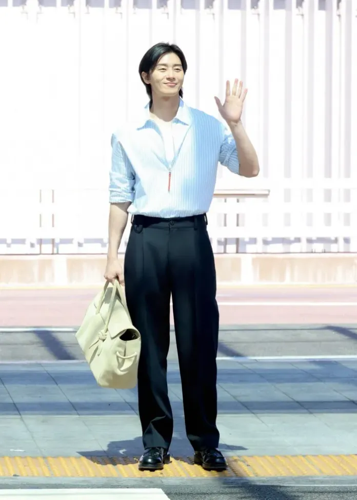 Park Seo-joon standing on a sidewalk, waving and smiling, wearing a light blue striped shirt and black trousers, holding a beige bag.