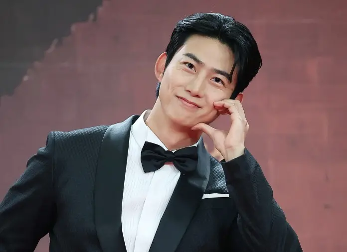 Taecyeon in a tuxedo making a playful gesture with his hand while smiling at the camera.