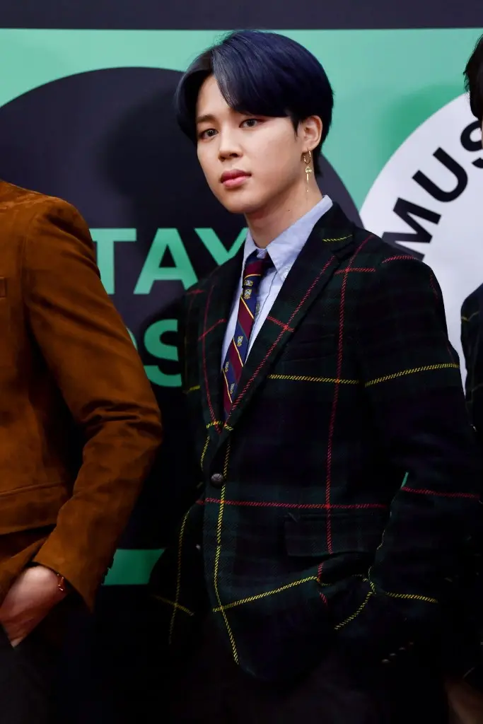Jimin with dark blue hair, wearing a plaid blazer over a light blue shirt and tie, standing confidently with his hand in his pocket.