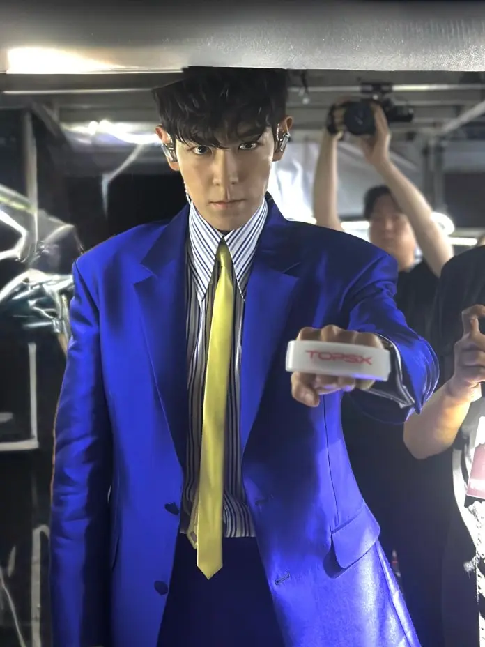 T.O.P in a striking blue suit with a yellow tie, holding a device labeled 'TOPSX' and looking intently at the camera. The background features blurred figures, indicating a lively environment.