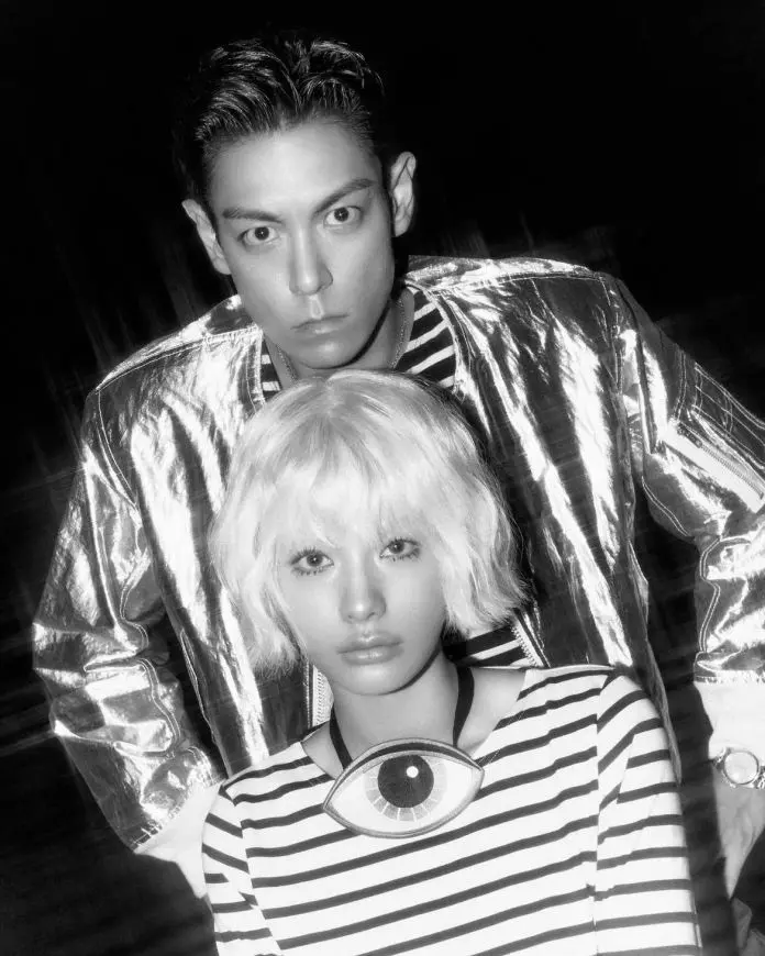A black and white photo featuring a T.O.P in a shiny silver jacket and a NANA with short, wavy blonde hair wearing a striped shirt with an eye-shaped pendant.