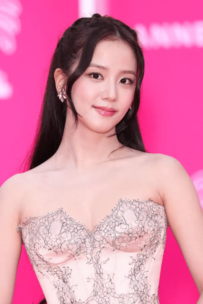 Jisoo with long black hair styled in waves, wearing a strapless, embellished pink gown. She is smiling and posed against a vibrant pink background.