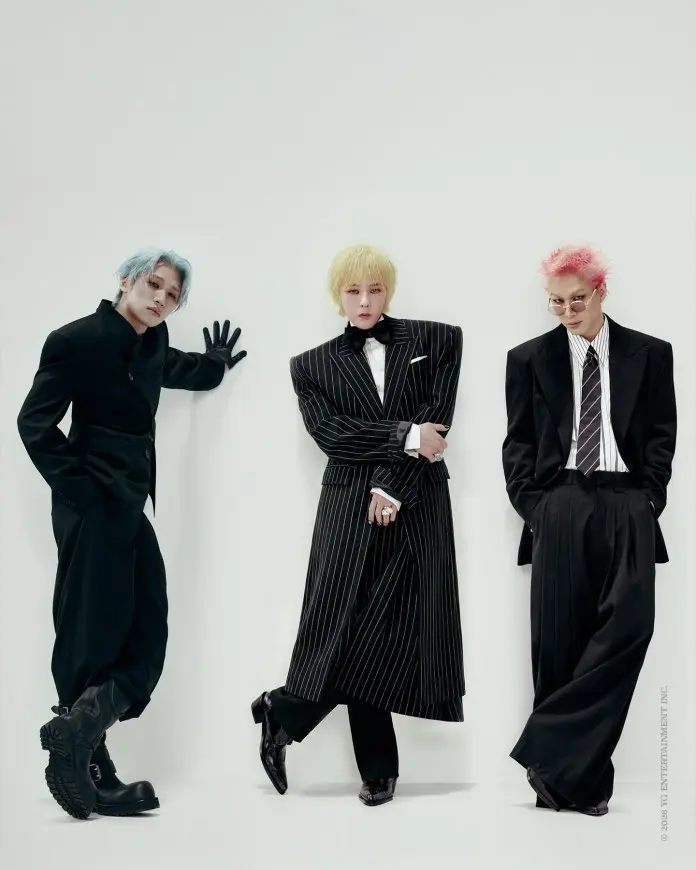 BIGBANG members posing against a plain white background, showcasing unique fashion styles. The person on the left has blue hair and wears a black outfit with bold boots, the center person has yellow hair and a striking pinstripe suit with a bow tie, and the person on the right has pink hair, glasses, and a classic black suit with a striped tie.