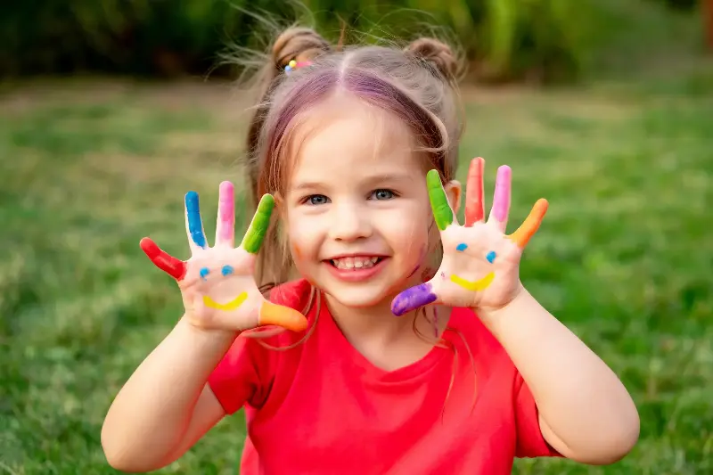 A small child girl with painted fingers in bright colors on her hands in summer on a green lawn.