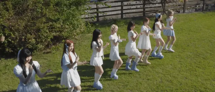 UNIS wearing matching white outfits, dancing in a row on a grassy area outdoors. The background includes greenery and a wooden fence.
