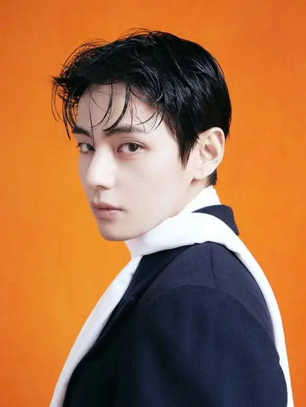 V with slicked-back dark hair, wearing a black jacket and white scarf, set against a bright orange background.