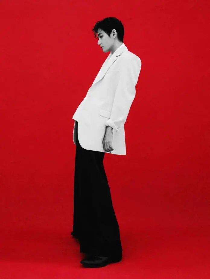 V in profile wearing a white blazer and black pants stands against a bright red background.