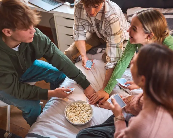 Four friends sitting on a bed playing a card game, with a bowl of popcorn in the center.
