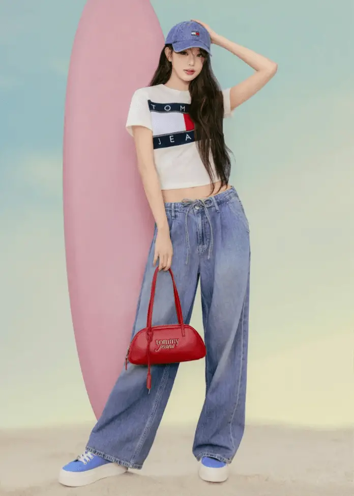 Jang Wonyoung stands confidently beside a pink surfboard, wearing a white cropped T-shirt with a blue and red logo, loose denim jeans, and blue sneakers. She holds a small red handbag and wears a matching blue cap.