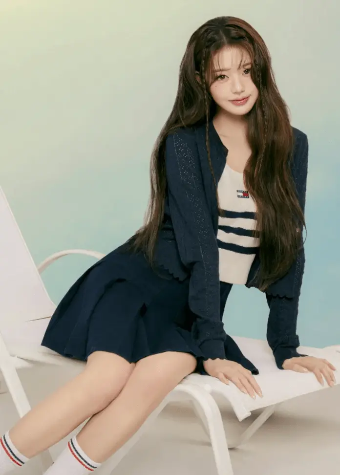 Jang Wonyoung sitting on a white lounge chair, wearing a navy cardigan over a striped top and a navy skirt. She has long, wavy hair and is posed casually with a relaxed expression against a soft, light-colored background.