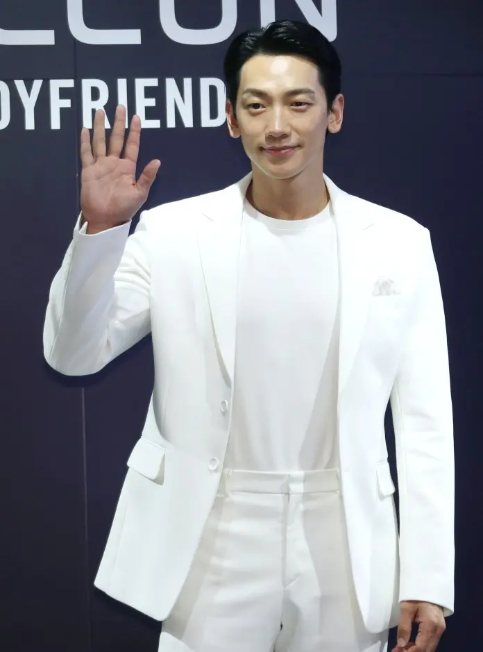Rain in a white suit and shirt waves, smiling at the camera, against a dark background with text.