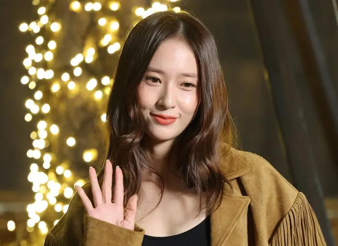 Krystal Jung with long, wavy hair wearing a brown suede jacket, smiling and waving in front of a backdrop of soft glowing lights.