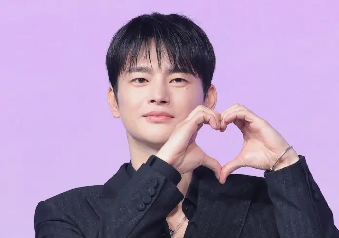 Seo In-guk  in a black suit making a heart shape with his hands against a purple background.