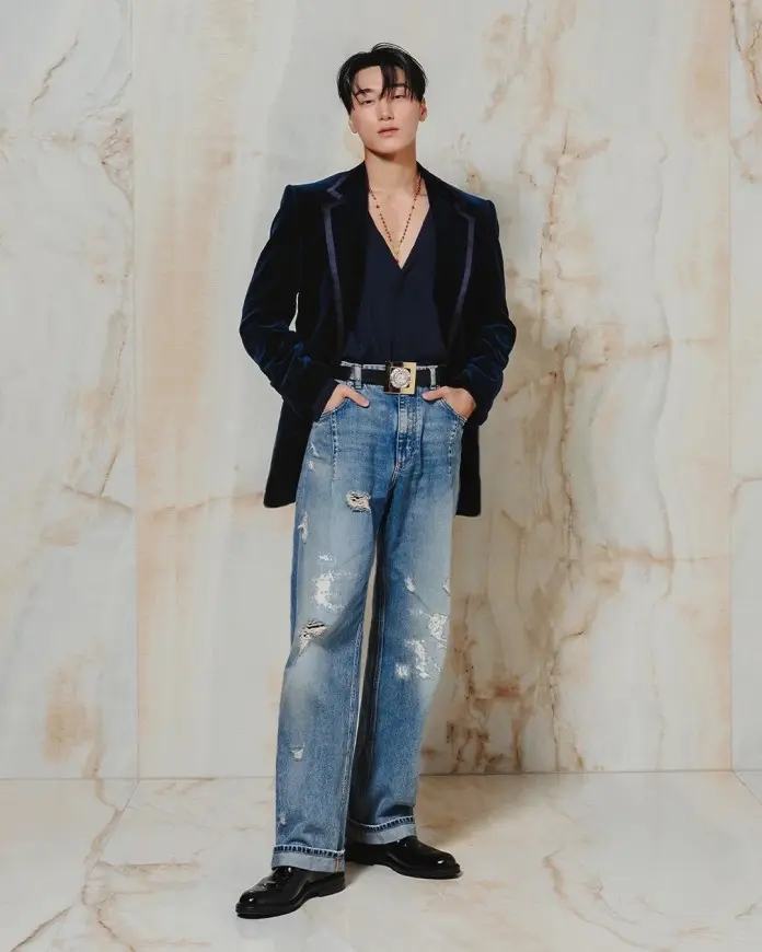 San poses confidently against a marble backdrop, wearing a dark velvet blazer over a deep v-neck shirt and distressed jeans, complemented by stylish black shoes and accessories.