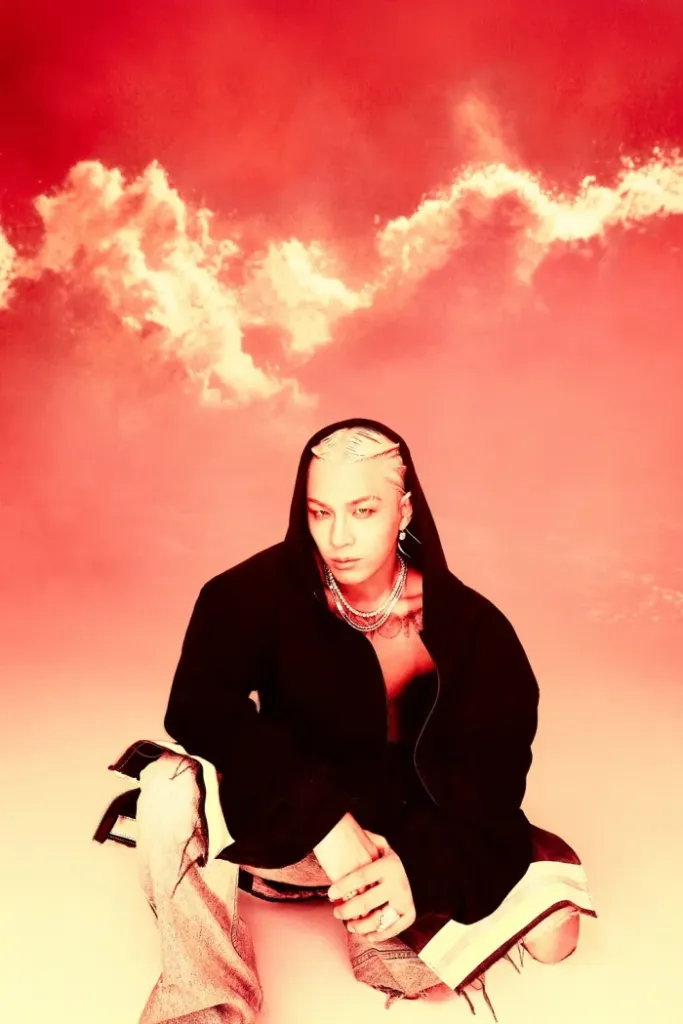 Taeyang sitting on the ground, dressed in a hooded black jacket and distressed jeans, against a vibrant red background with swirling clouds.