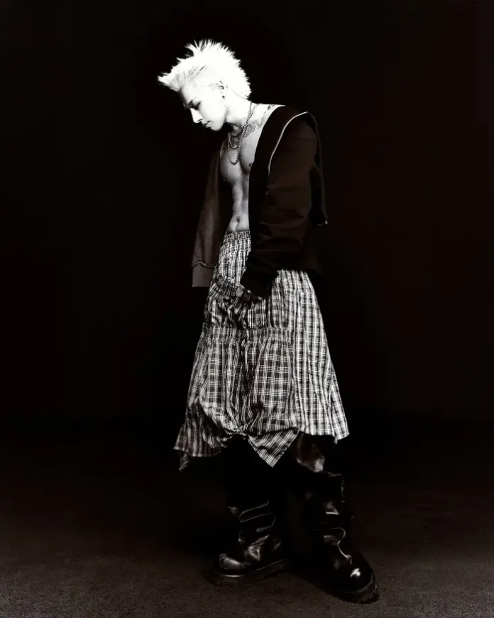 Taeyang with spiky white hair wearing a cropped black hoodie and a plaid skirt, standing in profile against a dark background, exuding an edgy fashion vibe.