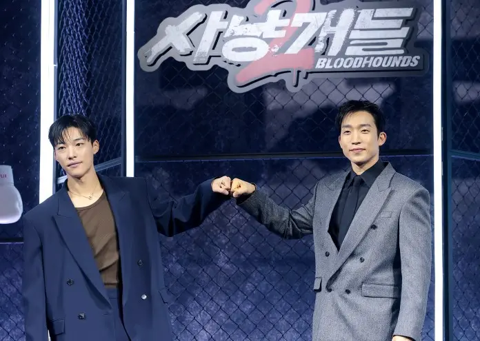 Lee Sang-yi posing together at a promotional event for 'Bloodhounds 2,' demonstrating a fist bump gesture in front of a backdrop featuring the show's logo.