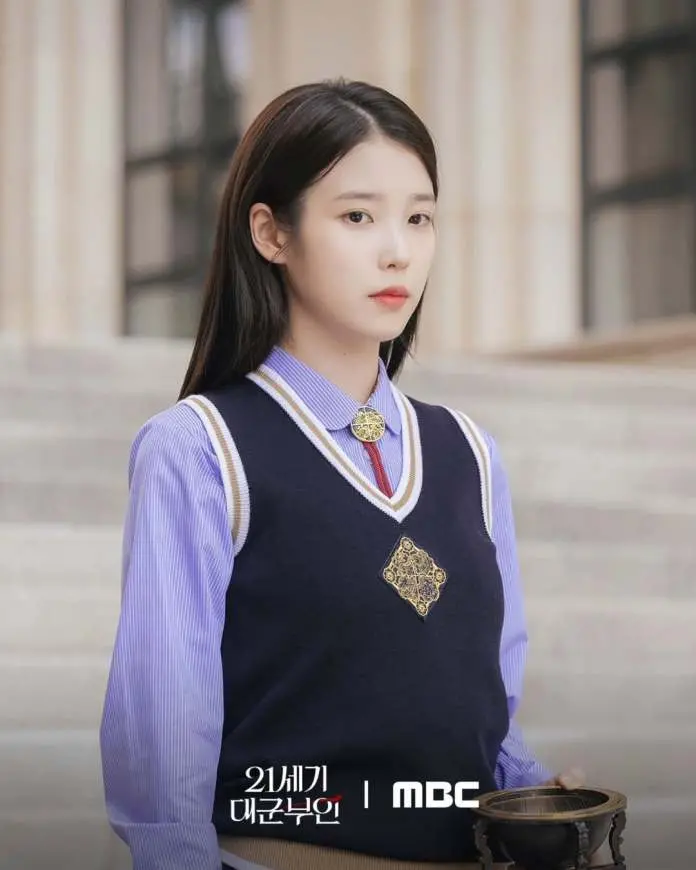 IU with long, dark hair is standing outdoors, wearing a navy blue vest over a light blue shirt. She holds a decorative bowl and looks serious, standing in front of stairs and a building.
