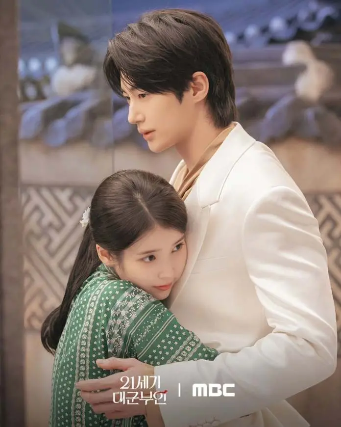 IU wearing a green patterned dress hugs Byeon Woo-seok in a white suit, both looking serene in a softly lit setting.