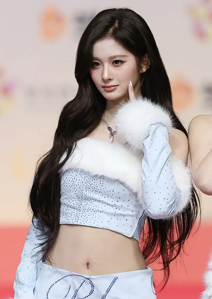 Sullyoon with long dark hair, wearing a sparkling light blue crop top with fur accents, flashes a playful gesture with her fingers while posing confidently at an event.