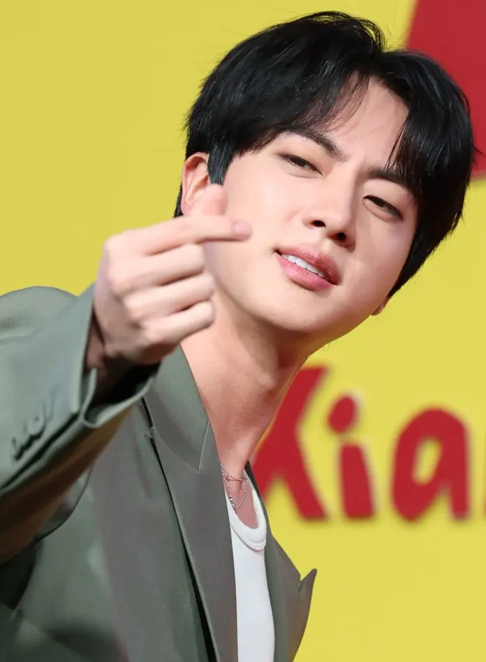 Jin with dark hair, wearing a stylish jacket, making a hand gesture toward the camera against a bright yellow background.