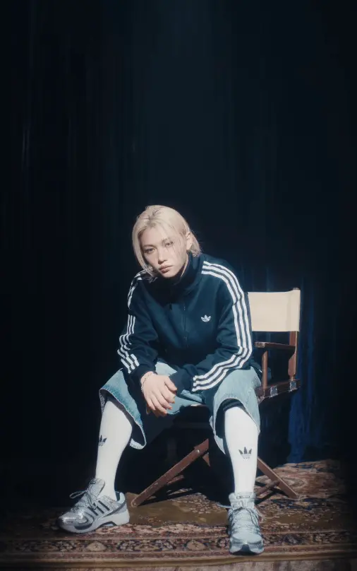 Felix sitting on a chair wearing an Adidas tracksuit, knee-length denim shorts, and sneakers, with a contemplative expression, set against a dark background.