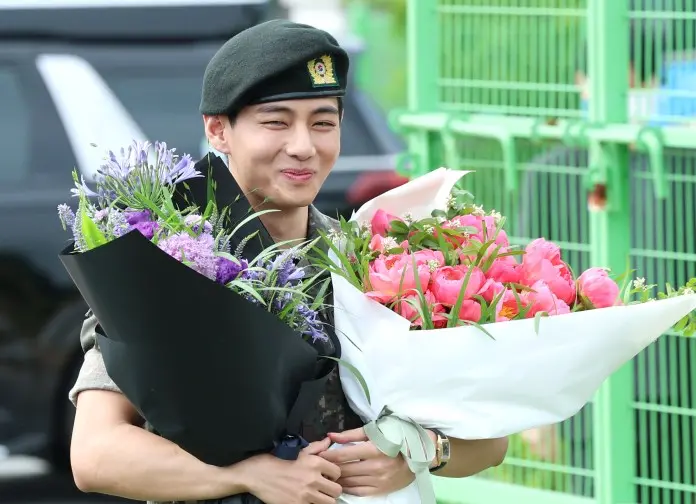 V in a military uniform and beret happily holds two large bouquets of flowers, one with pink peonies and the other with purple blooms.