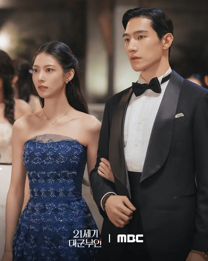 Queen and Prime Minister standing together at a formal event. The woman is wearing a blue lace strapless gown, while the man is dressed in a black tuxedo with a bow tie. They appear to be attending a high-class gathering.