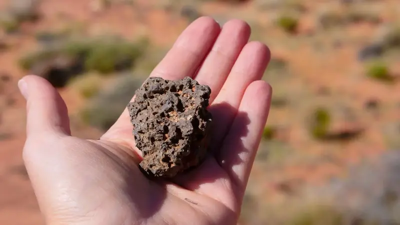 A hand holding a rough dark rock in a desert, evoking a meteorite recovered from the Sahara.