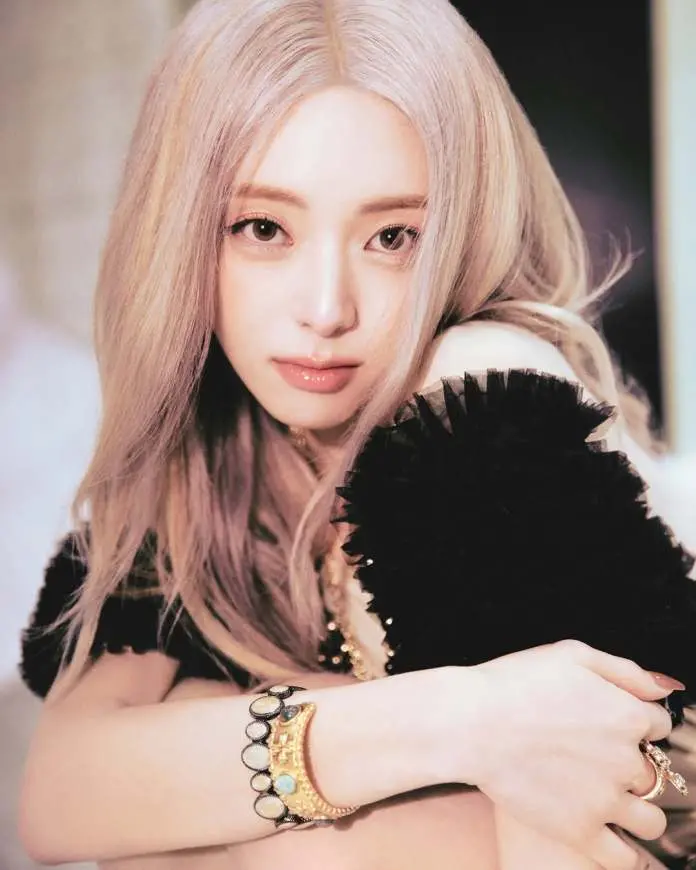 A close-up portrait of Yuna with long, light-colored hair, wearing a black, fluffy top, showcasing a gold bracelet on one wrist.