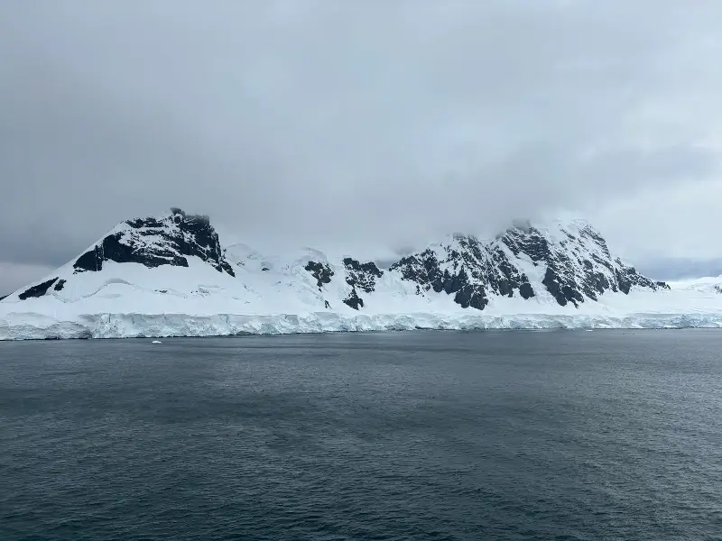 Snow-covered mountains and icebergs in Antarctica under a cloudy sky.