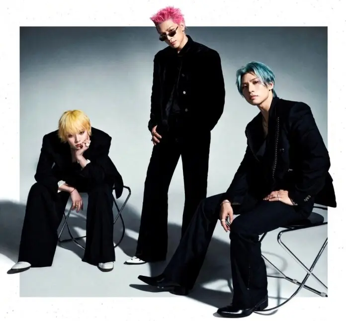 BIGBANG posing in stylish black outfits with colorful hair: one with pink hair standing, one with yellow hair sitting, and one with blue hair sitting.