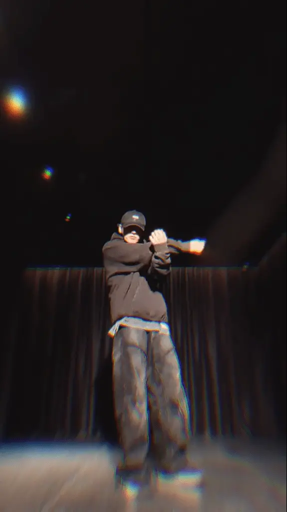 Jungkook performing on stage, wearing a black hoodie and cap, with a dramatic pose against a dark background.