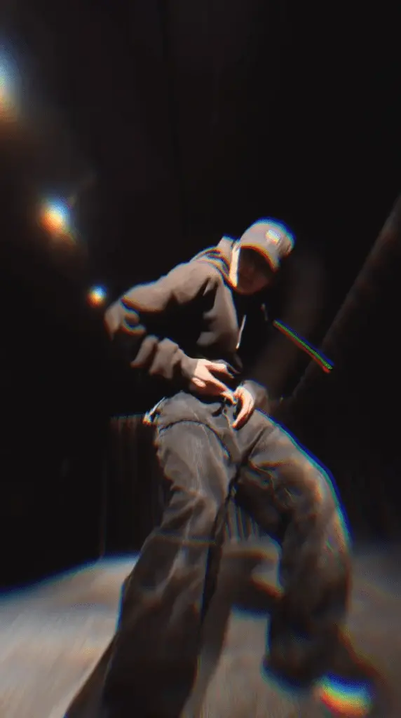 Jungkook in a dark space wearing a hoodie and cap, performing a pose with hands on their waist.