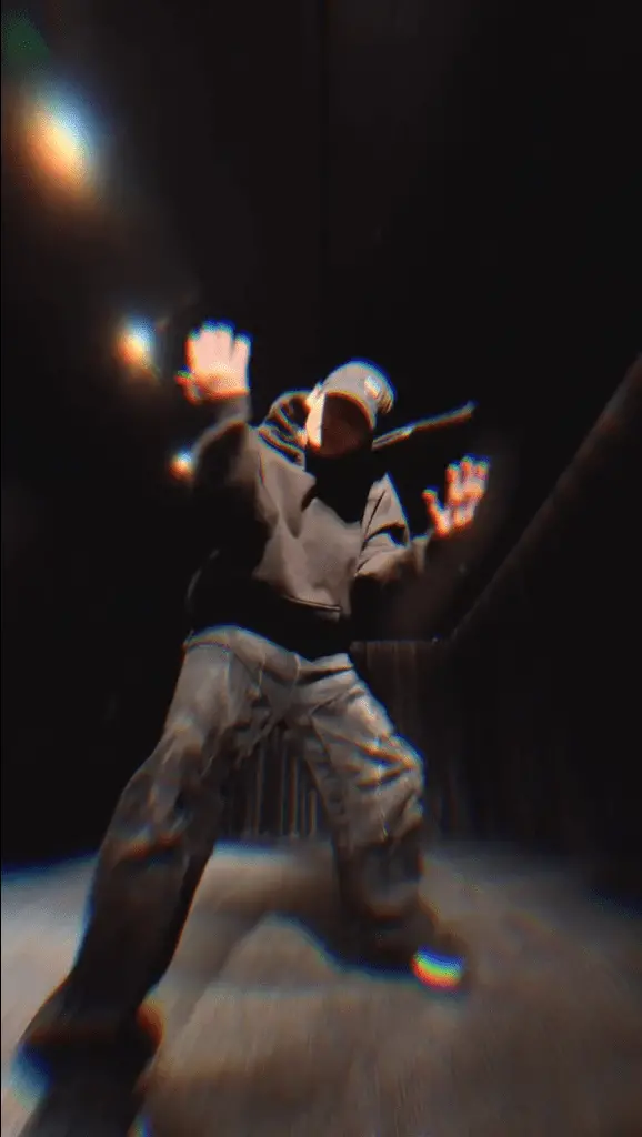 Jungkook wearing a black hoodie and cap is striking a dynamic pose in a dark room, with dramatic lighting effects highlighting their hands.