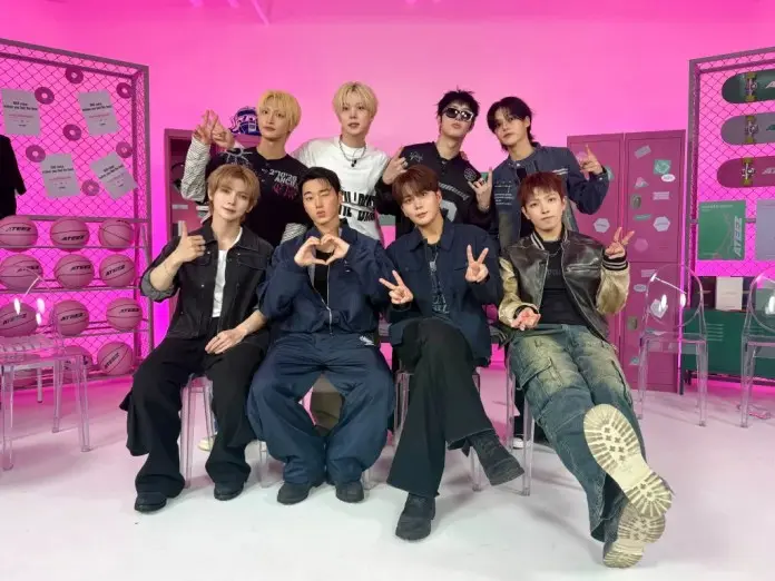 ATEEZ posing together in a brightly lit room with a pink background, sporting casual stylish outfits and various hand gestures, including peace signs and heart shapes.