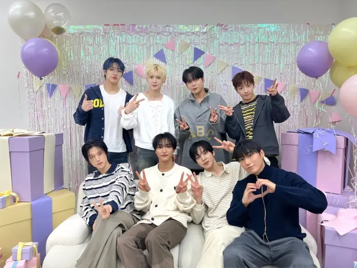 ATEEZ posing together in a festive setting, surrounded by balloons and decorative banners. They are seated and standing in two rows, smiling and making peace signs. Colorful gift boxes are placed nearby.