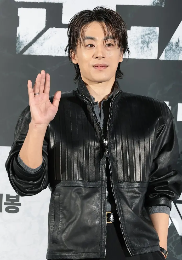 Koo Kyo-hwan wearing a black leather jacket and greeting with a hand wave at a press event.