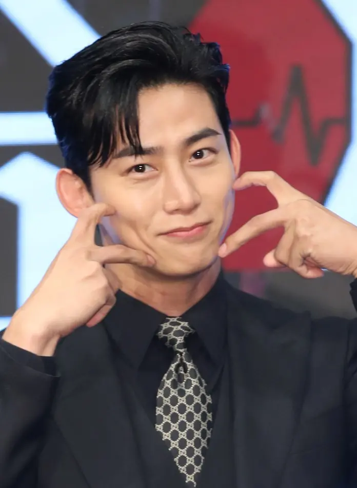 Ok Taecyeon in a dark suit and patterned tie is playfully making a gesture with his hands to his face, showcasing a cheerful expression.