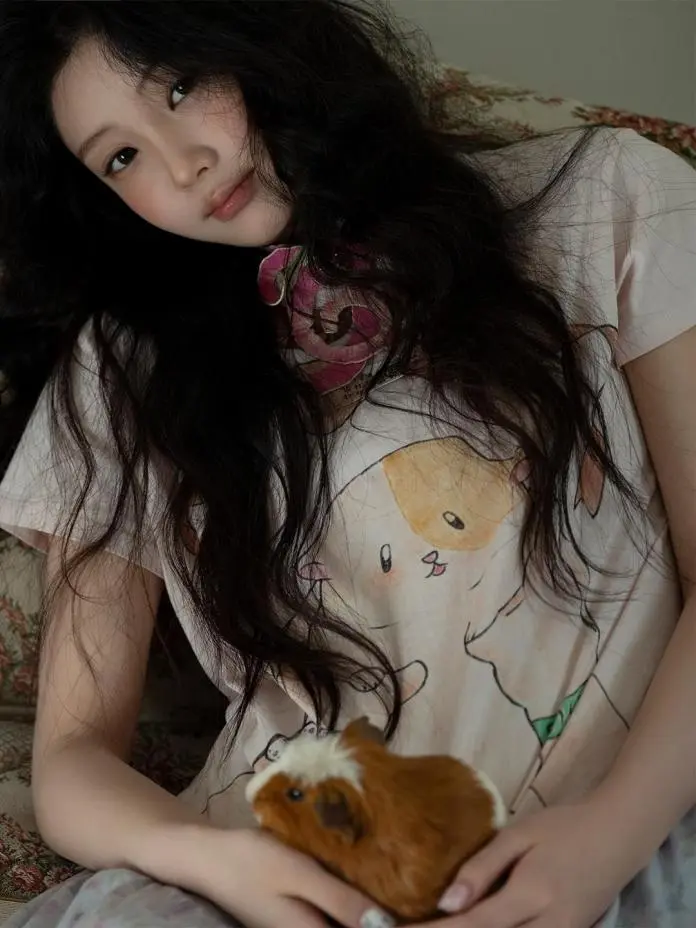 A young person with long dark hair, sitting on a floral couch, wearing a light-colored t-shirt with animal illustrations, holding a brown and white guinea pig.